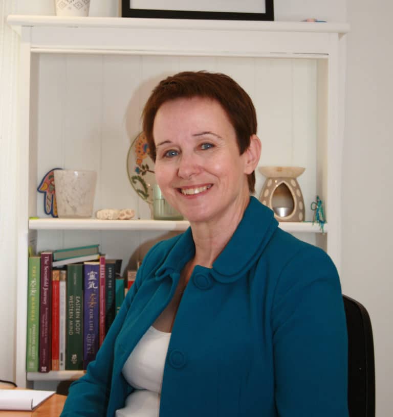 Image of Carol in a blue jacket, smiling sat in front of a white bookcase.
