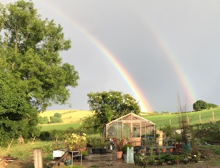 Image of a double rainbow over a greenhouse with trees and a hillside in the distance